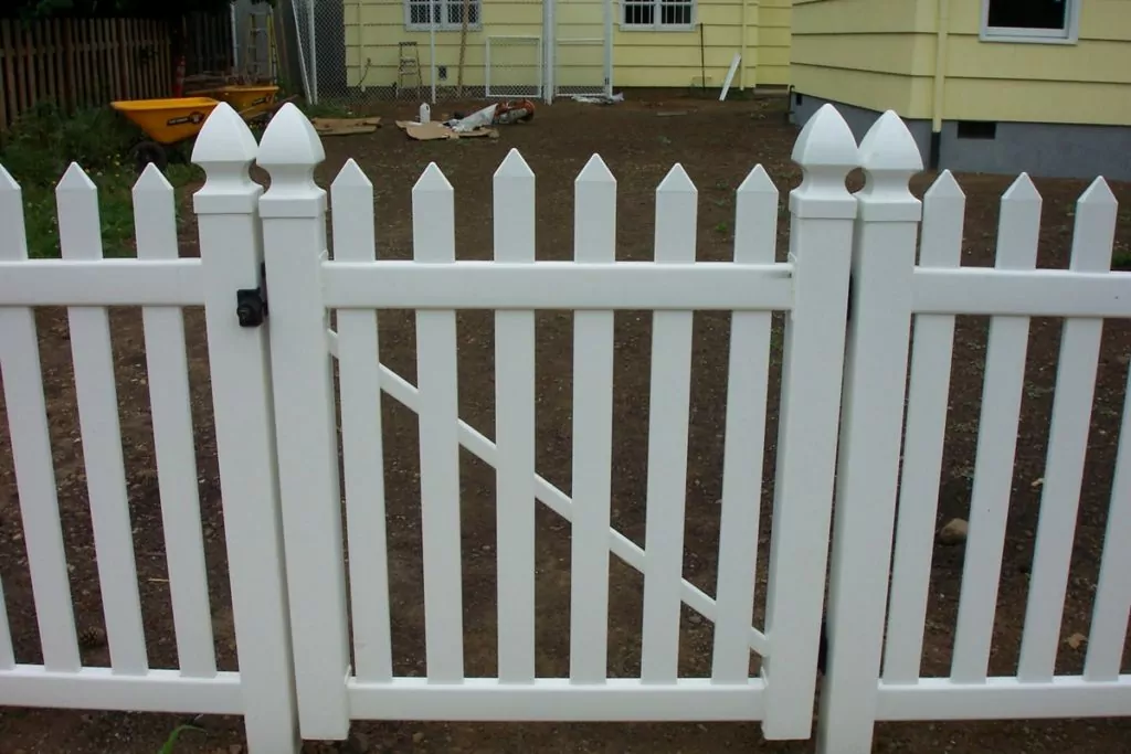 A white, vinyl picket fence gate in front of a yellow house to help illustrate fence post types